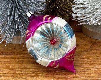 May include: A vintage-style glass Christmas ornament with a teardrop shape, featuring a vibrant magenta finish, a central silver indented reflector, and white painted accents, positioned in front of three small bottle brush trees.