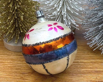 May include: A spherical glass Christmas ornament featuring a cream-colored base with hand-painted pink floral accents, a central horizontal band of metallic orange and dark blue, and vertical glittered lines extending from the center.