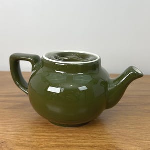 May include: A small, olive green ceramic teapot with a rounded body, a curved spout, and a square handle. The rim of the teapot is white. The teapot is sitting on a wooden surface.