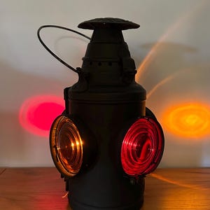 May include: A black vintage lantern with three colored lenses: amber and red. The lantern has a handle and a domed top. The light from the lenses casts red and orange circles on the background.