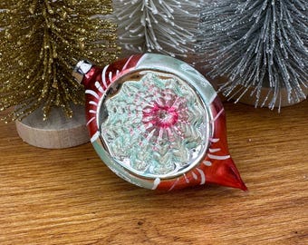 May include: A red and silver teardrop-shaped glass ornament featuring a central indented reflector with a pink and white floral pattern, positioned in front of three miniature bottle brush trees in gold, white, and silver.