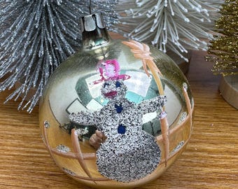 May include: A spherical glass Christmas ornament with a metallic gold finish, featuring a textured silver glitter snowman wearing a pink hat. The snowman is positioned behind a painted tan fence design, with a small metal cap at the top.