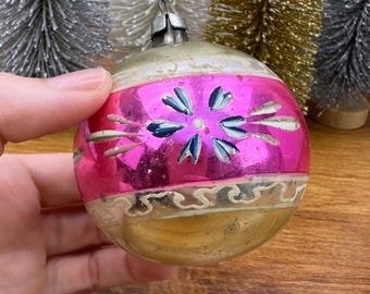 May include: A spherical glass Christmas ornament featuring a wide, reflective hot pink horizontal band decorated with a hand-painted blue floral motif and white accents, bordered by aged gold-toned glass sections and a silver metal cap.