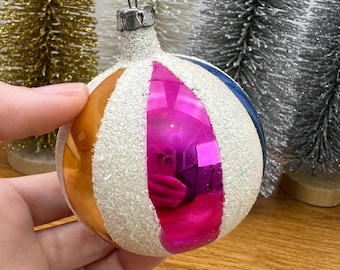 May include: A spherical glass Christmas ornament featuring alternating vertical panels of reflective orange, hot pink, and blue, separated by textured white glitter stripes. The ornament has a silver metal cap at the top for hanging.