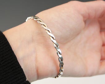 May include: A silver-colored braided bracelet. The bracelet is made of intertwined metal strands, creating a textured, woven appearance. The bracelet is worn on a wrist, with a black sleeve visible in the image.