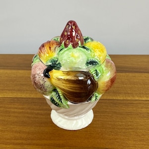 May include: A ceramic fruit-shaped jar with a lid. The jar is white with a pink base and is adorned with a variety of colorful fruits, including strawberries, apples, and pears. The lid is topped with a large red strawberry.