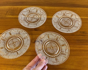 May include: Four clear glass saucers with an embossed floral design are arranged on a wooden surface. The saucers have a circular shape with a detailed pattern around the edges and center. One saucer is being held by a hand.