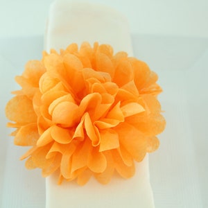 Puede incluir: Una gran flor de papel naranja brillante, tambi&eacute;n conocida como pompon de papel de seda, se coloca sobre una servilleta blanca.