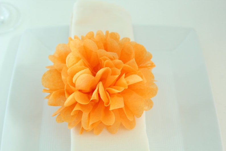 Puede incluir: Una gran flor de papel naranja brillante, tambi&eacute;n conocida como pompon de papel de seda, se coloca sobre una servilleta blanca.