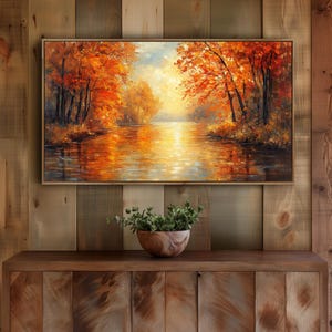 Könnte beinhalten: Ein gerahmtes Landschaftsgemälde zeigt eine herbstliche Szene. Das Kunstwerk zeigt einen Fluss, der die leuchtenden Farben des Herbstlaubes widerspiegelt, mit Bäumen in Orange- und Rottönen. Das Gemälde wird über einem Holzschrank ausgestellt.