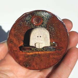 May include: A handmade ceramic pendant with a unique design. The pendant features a circular, reddish-brown base with a small, off-white figure in a recessed area. The figure has two small dots for eyes and a rectangular shape below. A small hole is at the top.