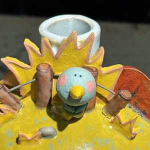 May include: A hand-painted ceramic decorative container. The container is round and yellow with a sunburst design around the top. A small blue bird with pink cheeks is perched on the top. The container has a small opening at the top.