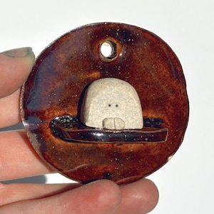 May include: A handmade ceramic pendant with a brown glaze. The pendant features a small, off-white, rounded figure with two dots and a line, possibly a face, inside a boat-like shape. A hole is at the top for hanging.