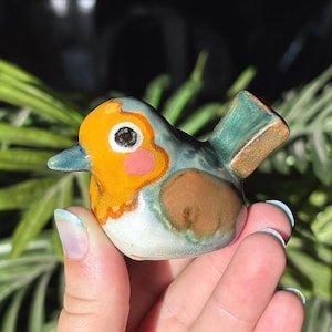 May include: A small ceramic bird figurine featuring a glossy finish with a bright orange face, a pink cheek patch, and a dark eye. The body is a mix of light blue and green tones with a brown wing patch and a pointed tail. The beak is blue.