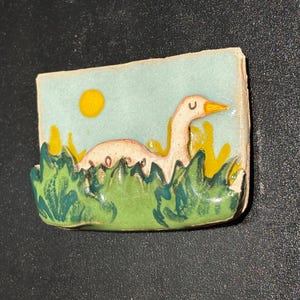 May include: A ceramic magnet featuring a goose swimming in a pond. The magnet has a light blue background with a yellow sun. The goose is white with an orange beak, and the grass is green and yellow.