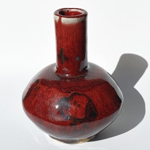 May include: A small, round, ceramic vase with a deep red glaze. The vase has a narrow neck and a wide, rounded body. The glaze has dark, mottled areas, adding depth and visual interest. The interior of the neck is a lighter color.