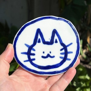 May include: A white ceramic coaster with a hand-drawn blue cat face design. The cat has a simple, stylized face with pointed ears, whiskers, and a small smile. The coaster has a blue border and is held in a hand.