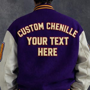 Op de afbeelding: Paarse en crèmekleurige varsityjas met de tekst "CUSTOM CHENILLE YOUR TEXT HERE" in gouden letters. De jas heeft crèmekleurige mouwen en goudgele afwerking rond de kraag.