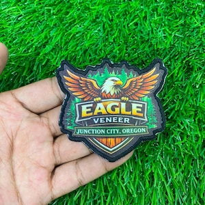 May include: A shield-shaped embroidered patch featuring a bald eagle with spread wings above a forest background. The text EAGLE VENEER is printed in yellow and white, with JUNCTION CITY, OREGON written in white on a green banner at the bottom.