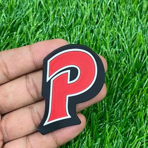 May include: A flat, stylized letter P emblem featuring a bold red center, a thin white outline, and a thick black border. The design has a sharp, angular typeface with a modern aesthetic, presented as a solid, smooth-surfaced decorative patch.