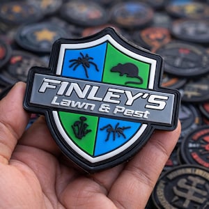Può includere: Una toppa a forma di scudo con la scritta "FINLEY'S Lawn & Pest" in bianco e grigio. La toppa presenta un design blu e verde con illustrazioni di insetti e roditori. Altre toppe sono sullo sfondo.