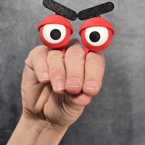 May include: Two red, cartoon eye rings with black eyebrows are held up by a hand. The eyes have white irises and black pupils. The background is a mottled grey.