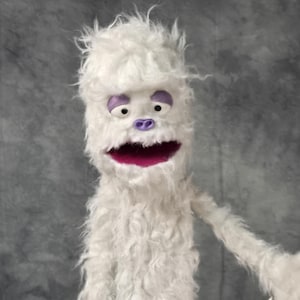 May include: A white puppet with a shaggy, textured body and head. The puppet has purple eye accents, a purple nose, and a wide, open mouth with a magenta interior. The puppet's arms are extended, and it appears to be made of a soft, plush material.
