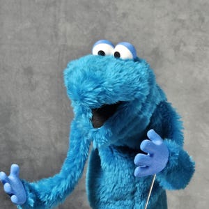 Könnte beinhalten: Eine blaue Krümelmonster-Puppe mit großen weißen und blauen Augen, einem schwarzen Mund und blauen Händen. Die Puppe besteht aus einem weichen, pelzigen Material und ist für die Bedienung mit Stäben konzipiert.