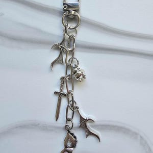 May include: Silver-coloured keychain with a lobster clasp and chain. The chain holds several charms, including a sword, a masquerade mask, a rose, and two bat wing charms. The background is a white and grey marble pattern.