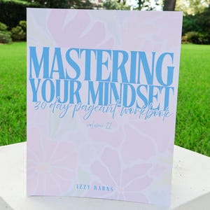 May include: A softcover workbook titled Mastering Your Mindset 30 day pageant workbook volume II by Izzy Karns. The cover features a light pink background with a subtle white floral pattern and bold blue text for the main title.