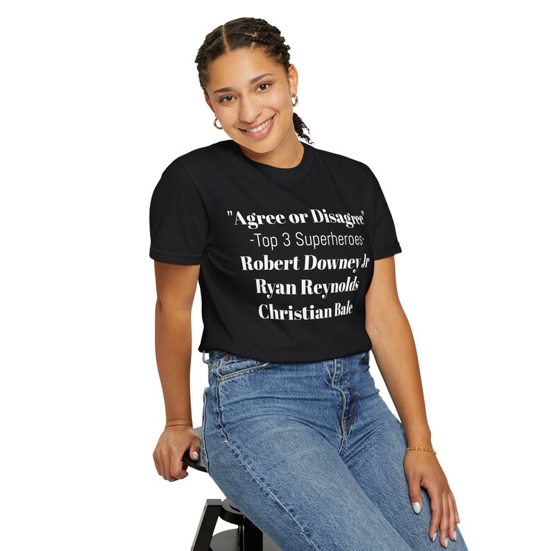 A smiling woman wearing a black t-shirt with white text and blue jeans is sitting on a stool.