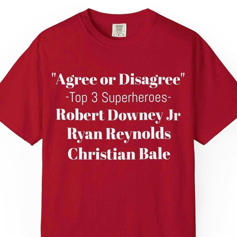A red t-shirt with white text that reads "Agree or Disagree" and "Top 3 Superheroes" along with the names Robert Downey Jr, Ryan Reynolds, and Christian Bale.