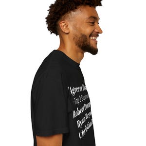 A man with curly hair and a beard is smiling and looking to the side. He is wearing a black t-shirt with white text on it.