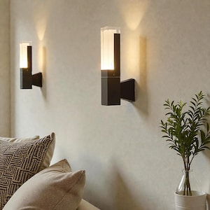 May include: Two modern, black wall sconces with rectangular, frosted glass shades are mounted on a light-colored wall. The sconces are illuminated, casting a warm glow. A vase with greenery and decorative pillows are in the foreground.