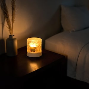 Puede incluir: Una lámpara cilíndrica texturizada y encendida, con base blanca, está sobre una mesita de noche de madera oscura. La lámpara emite un brillo amarillo cálido. Un jarrón gris con plantas secas está a la izquierda de la lámpara. Una cama con sábanas blancas está al fondo.