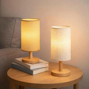 Puede incluir: Dos lámparas de mesa cilíndricas con pantallas de tela beige y bases de madera. Una lámpara está sobre una pila de libros, la otra sobre una mesa redonda de madera. Las lámparas emiten una luz cálida, ideal para un dormitorio o salón.