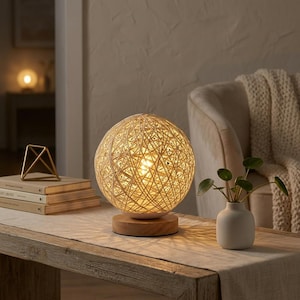 May include: A spherical table lamp with a woven, cream-colored shade sits on a wooden base. The lamp is lit, casting a warm glow. The lamp is on a wooden table with a small white vase and a plant.