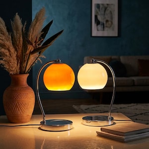 May include: Two modern table lamps with spherical shades. One lamp has an orange shade, the other a white shade. Both have a chrome base and arched support. A vase with dried plants and a stack of books are also visible.