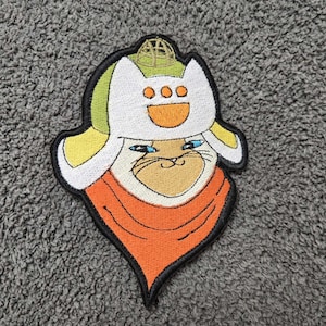 May include: Embroidered patch featuring a cartoon cat wearing a white hat with yellow and green accents. The cat has blue eyes and an orange scarf. The patch has a black border and is on a gray surface.