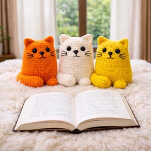 May include: Three crochet cat ornaments in orange, white, and yellow sit on a fluffy white surface. Each cat has black eyes and whiskers. An open book rests in front of the cats, creating a cosy, whimsical scene.