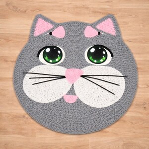 May include: A handmade, gray crocheted cat rug with pink accents. The cat's face features large green eyes, a pink nose, and white cheeks. Black whiskers and pink inner ears complete the design. The rug is round and sits on a wooden surface.