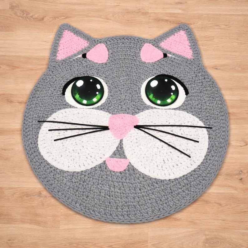 May include: A handmade, gray crocheted cat rug with pink accents. The cat's face features large green eyes, a pink nose, and white cheeks. Black whiskers and pink inner ears complete the design. The rug is round and sits on a wooden surface.