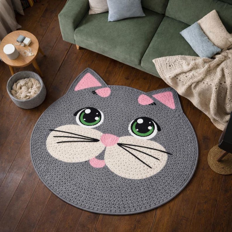 May include: A gray, cat-shaped rug with a white face, pink ears, and green eyes. Black whiskers and a pink nose complete the design. The rug is round and made of a woven material, perfect for a child's room or a playful space.