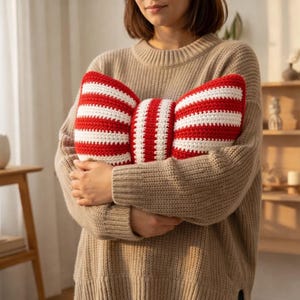 May include: A red and white striped bow-shaped crochet pillow. The pillow is held by a person wearing a tan sweater. The pillow's design features alternating red and white stripes, creating a visually appealing pattern. The crochet work is visible, adding texture.