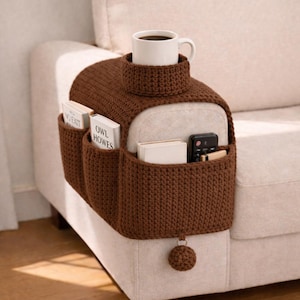 May include: A brown, crocheted sofa arm organizer with a built-in cup holder and multiple pockets. The organizer is designed to fit over the arm of a sofa and includes a small, decorative pom-pom. A white mug with coffee sits in the cup holder.