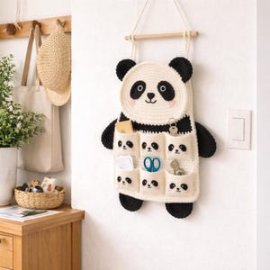 Può includere: Un organizer a forma di panda all'uncinetto fatto a mano, color crema con dettagli neri. L'appendino a parete a forma di panda presenta un volto sorridente, orecchie nere e più tasche con facce di panda. È appeso a un'asta di legno.