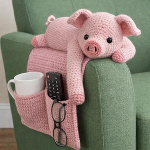 Puede incluir: Un organizador de reposabrazos en forma de cerdo rosa claro hecho a mano, con bolsillos. El organizador contiene una taza blanca, un mando a distancia y gafas. El cuerpo del cerdo se extiende sobre el reposabrazos de una silla verde.