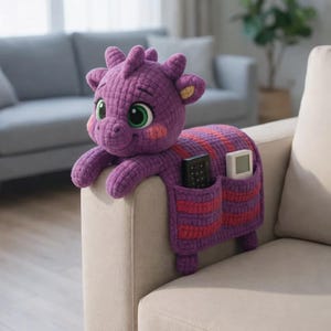 Puede incluir: Un organizador de reposabrazos de sofá con forma de dragón morado tejido a ganchillo. El dragón tiene grandes ojos verdes, mejillas rosadas y un cuerpo a rayas en morado y rojo. Dos bolsillos contienen un mando a distancia y un pequeño dispositivo blanco.