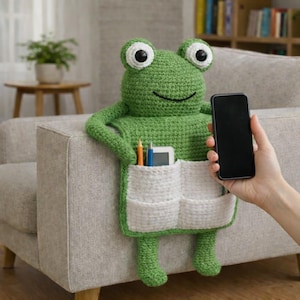 Op de afbeelding: Een groene gehaakte kikker-vormige organizer met witte zakken, ontworpen om over een bankarm te hangen. De kikker heeft grote witte ogen met zwarte pupillen en houdt een smartphone vast. Potloden en een kleine tablet zitten in de zakken.