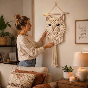 Puede incluir: Un colgante de pared de macramé color crema con ojos negros, nariz rosa y largos bigotes. La cabeza del gato está montada en una clavija de madera y tiene una larga borla. El colgante de pared se exhibe en una pared beige.